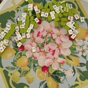 May include: A collection of green and white Mahjong tiles scattered on a tray. The tray features a floral design with pink blossoms, lemons, and green leaves. The tiles display various symbols and numbers.