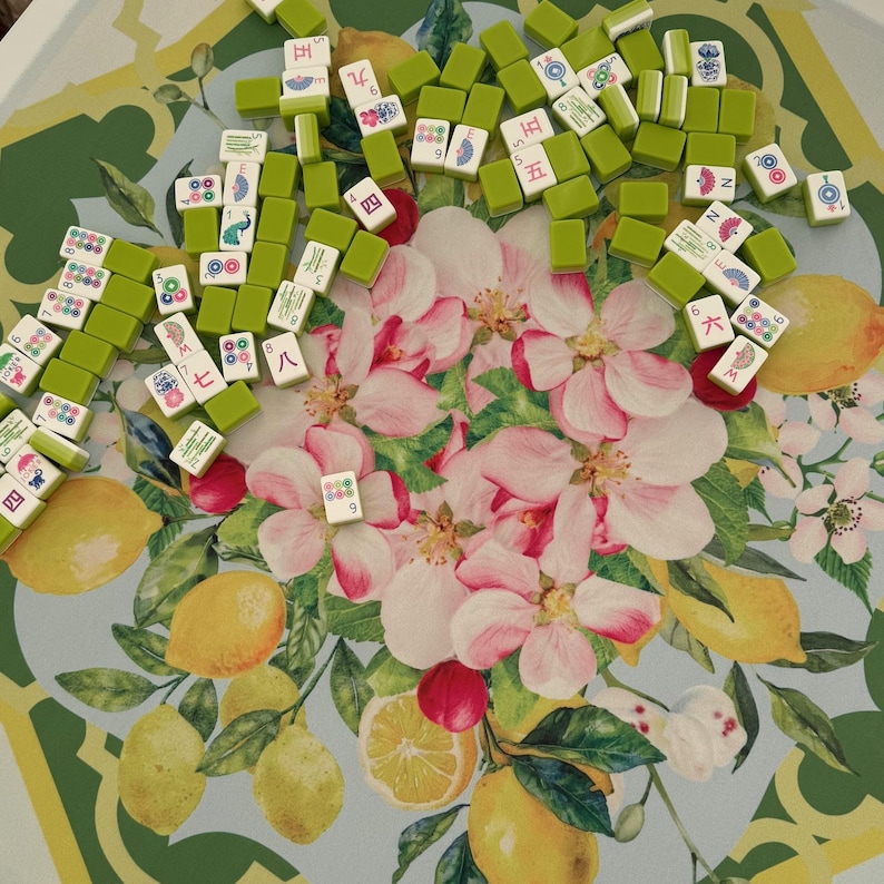 May include: A collection of green and white Mahjong tiles scattered on a tray. The tray features a floral design with pink blossoms, lemons, and green leaves. The tiles display various symbols and numbers.