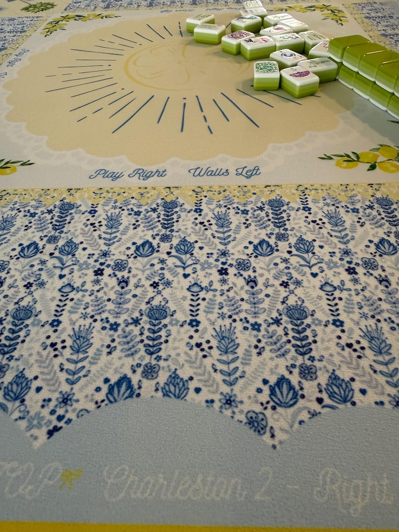 Puede incluir: Un juego de Mahjong sobre un tapete estampado. El tapete presenta un dise&ntilde;o de sol y patrones florales en azul y blanco. Las fichas verdes y blancas est&aacute;n apiladas sobre el tapete. El texto en el tapete dice "Play Right Walls Left" y "Charleston 2 - Right".