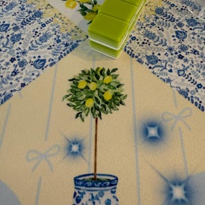 Puede incluir: Una alfombra decorativa con un dise&ntilde;o de limonero en una maceta azul y blanca. La alfombra tiene un fondo amarillo claro con detalles azules y un borde floral. Una pila de baldosas verdes descansa sobre la alfombra.