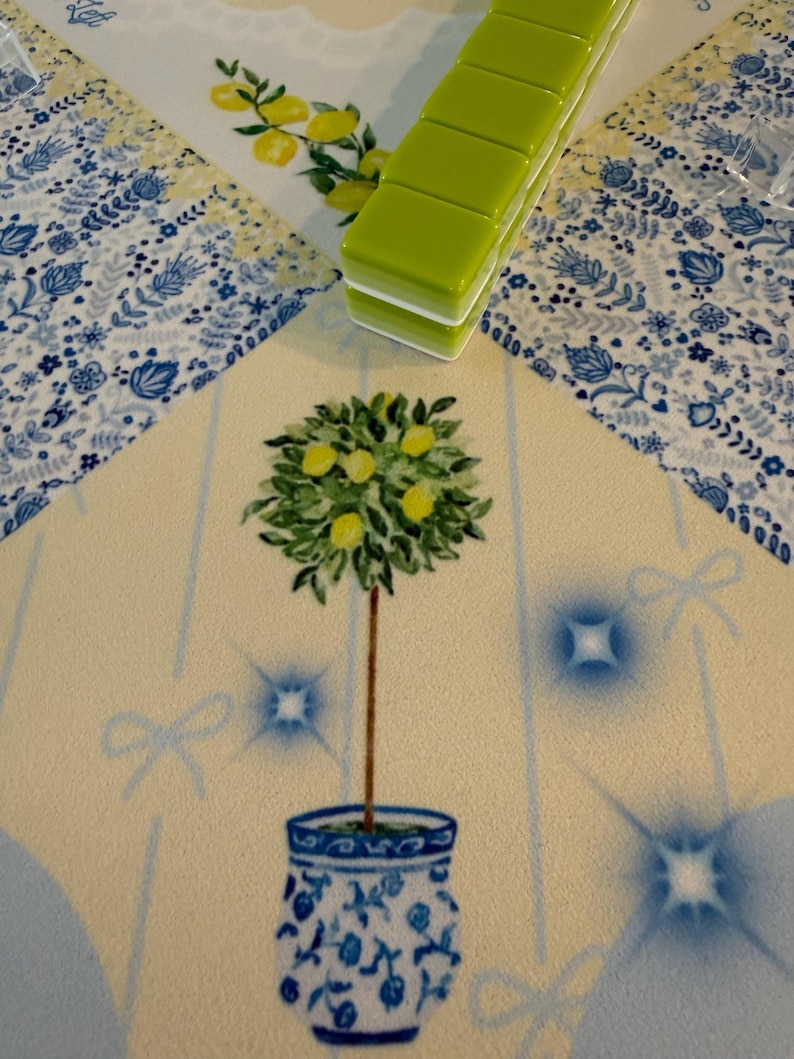 Puede incluir: Una alfombra decorativa con un dise&ntilde;o de limonero en una maceta azul y blanca. La alfombra tiene un fondo amarillo claro con detalles azules y un borde floral. Una pila de baldosas verdes descansa sobre la alfombra.