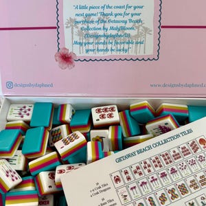 May include: A Mahjong tile set from the Getaway Beach Collection by Mahj Bloom. The box lid features text and palm tree silhouettes. Inside are white tiles with colorful beach-themed graphics and teal, pink, and yellow striped sides. A reference card is included.