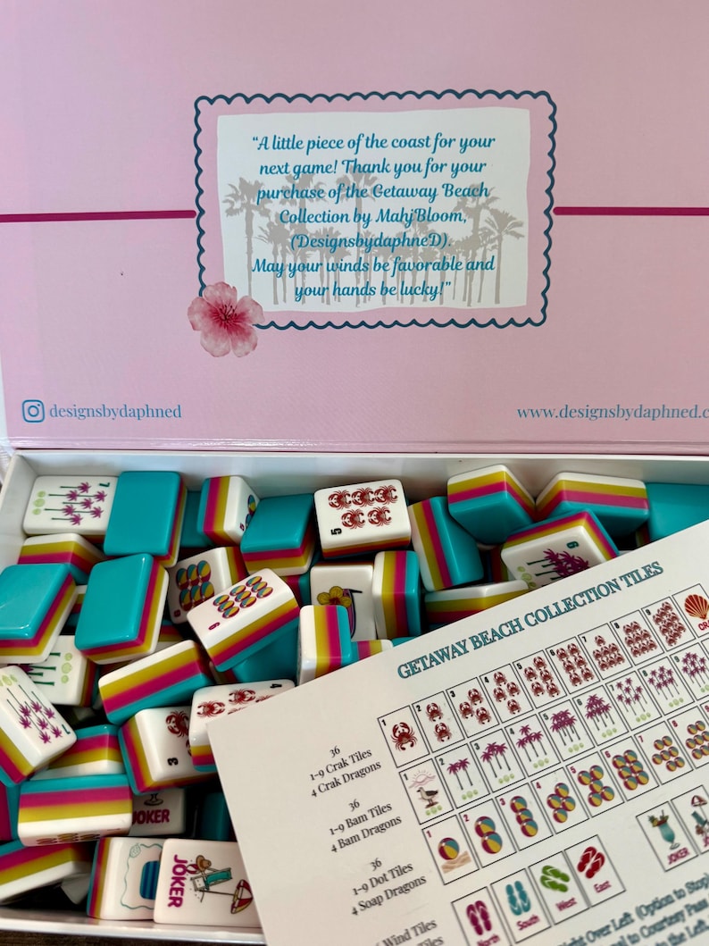 May include: A Mahjong tile set from the Getaway Beach Collection by Mahj Bloom. The box lid features text and palm tree silhouettes. Inside are white tiles with colorful beach-themed graphics and teal, pink, and yellow striped sides. A reference card is included.