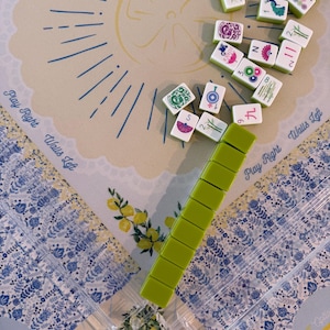 Puede incluir: Un juego de Mahjong con fichas verdes y un soporte para fichas verde claro. El juego est&aacute; dispuesto sobre un tapete estampado con las palabras "Play Right Walls Left". Las fichas son blancas con bordes verdes y varios s&iacute;mbolos y n&uacute;meros.
