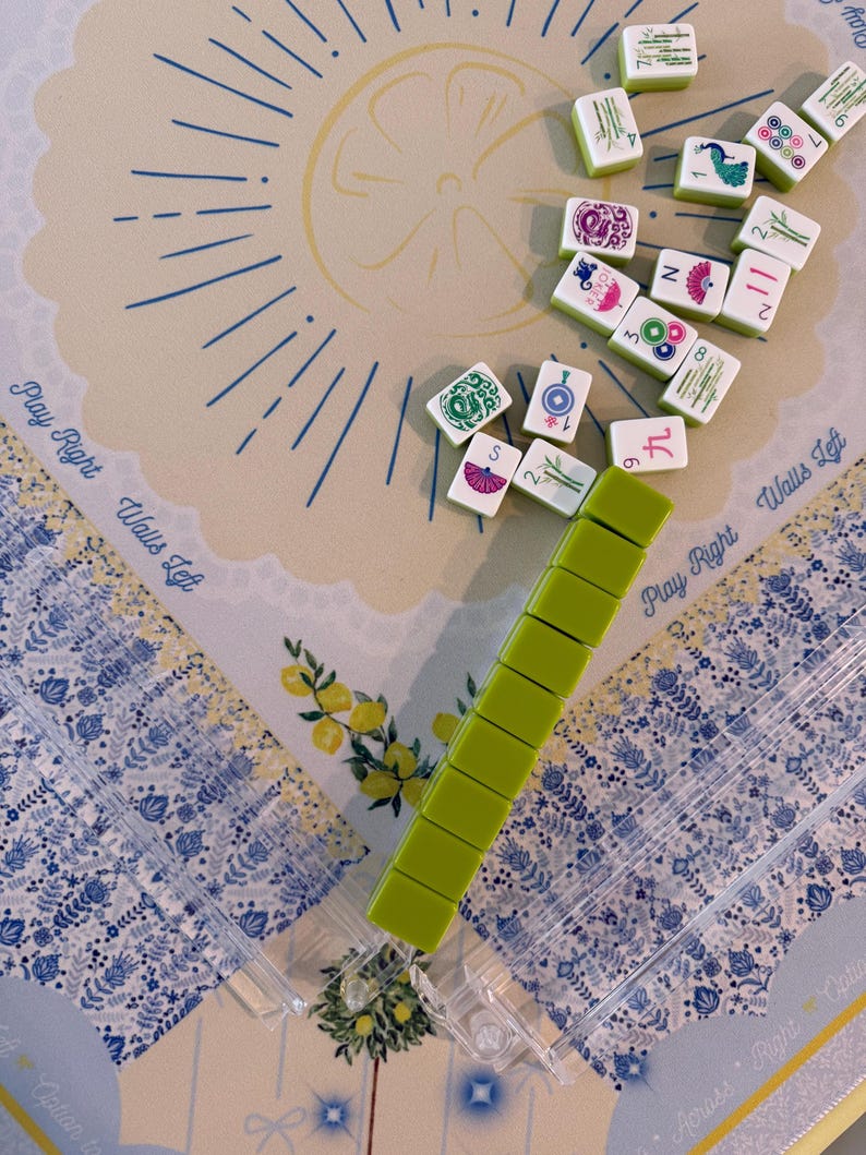 Puede incluir: Un juego de Mahjong con fichas verdes y un soporte para fichas verde claro. El juego est&aacute; dispuesto sobre un tapete estampado con las palabras "Play Right Walls Left". Las fichas son blancas con bordes verdes y varios s&iacute;mbolos y n&uacute;meros.