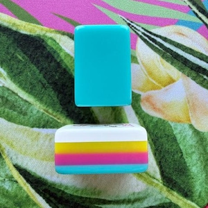 May include: Two rectangular mahjong tiles are displayed. The top tile is solid turquoise. The bottom tile features a white top section with a small black graphic, followed by horizontal stripes of yellow, magenta, and turquoise on its side.