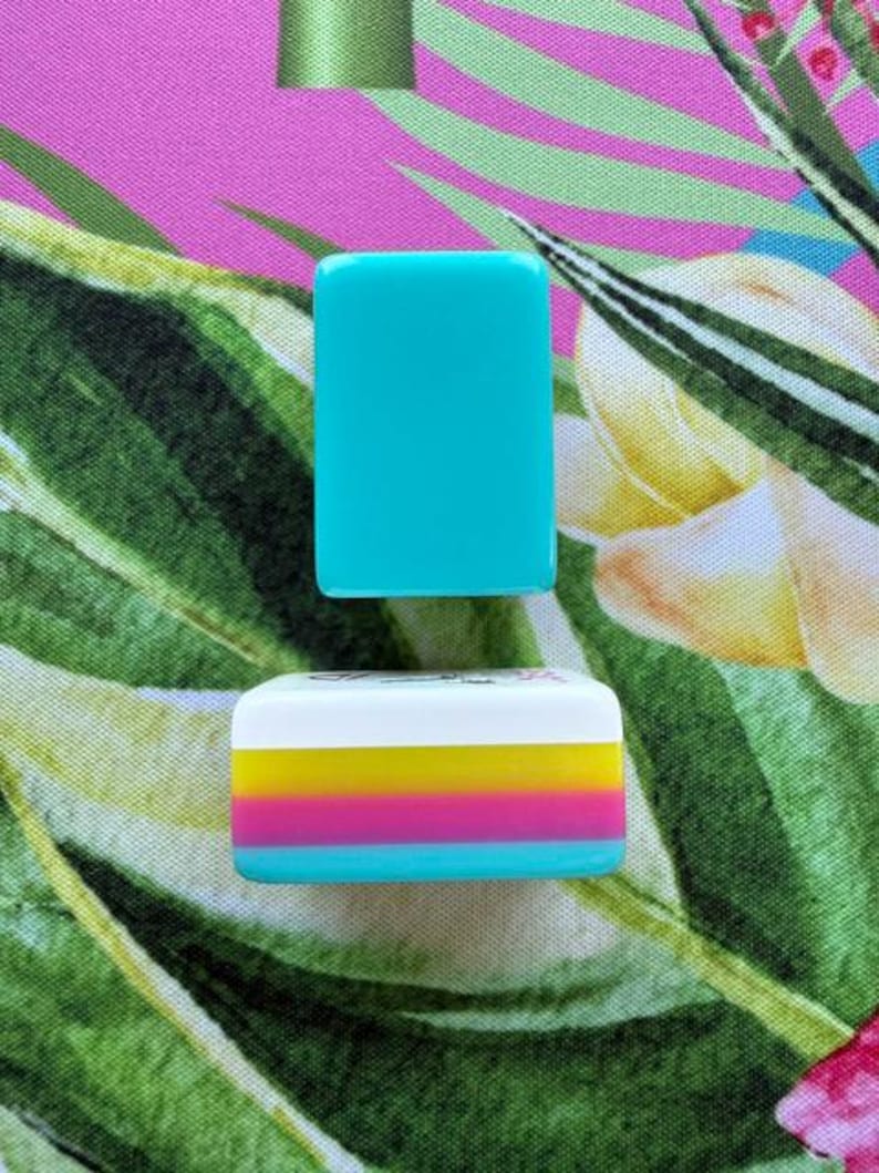 May include: Two rectangular mahjong tiles are displayed. The top tile is solid turquoise. The bottom tile features a white top section with a small black graphic, followed by horizontal stripes of yellow, magenta, and turquoise on its side.
