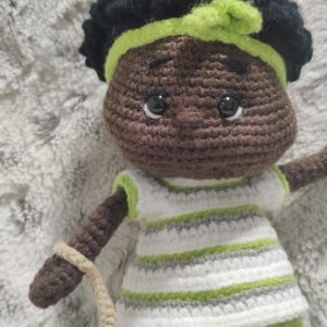May include: A handmade crochet doll with dark skin, black hair, and a green headband. The doll wears a white and green striped shirt and green shorts. It holds a beige crocheted strap and a maroon flower.