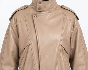 May include: A beige leather jacket featuring a high collar with two snap buttons, an asymmetrical front closure with a single snap button, shoulder epaulets, and long sleeves. The garment is constructed with visible seam detailing across the chest and shoulders, providing a structured appearance.