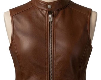 May include: A brown sleeveless vest featuring a stand-up collar with a single snap button closure. The garment includes a full-length silver-toned metal zipper down the center and two zippered pockets at the lower front, all constructed from smooth material.
