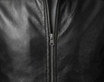 May include: A black jacket featuring a smooth, textured surface and a central vertical metal zipper. The garment includes a stand-up collar and visible stitching along the front panels, showcasing a clean, structured design with metallic hardware.