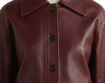 May include: A deep burgundy jacket with a smooth, leather-like finish, featuring a wide pointed collar and a front button placket with three dark, circular buttons. The garment has long sleeves and a structured, clean-lined design throughout.