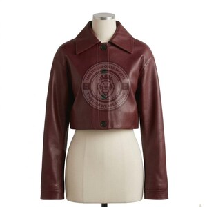 Puede incluir: Una chaqueta corta de cuero color burdeos con cuello en punta, mangas largas y cierre frontal de botones. El centro presenta un logotipo circular con un gr&aacute;fico de cabeza de le&oacute;n y el texto MASTER EMPOWER SPORTS TOGETHER WE ARE STRONG ESTD 2020.