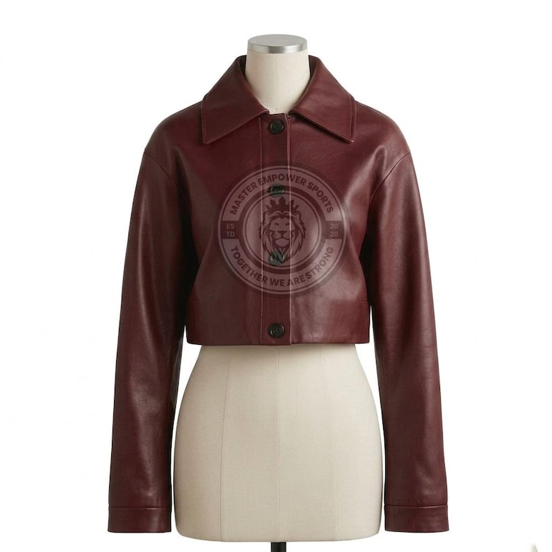 Puede incluir: Una chaqueta corta de cuero color burdeos con cuello en punta, mangas largas y cierre frontal de botones. El centro presenta un logotipo circular con un gr&aacute;fico de cabeza de le&oacute;n y el texto MASTER EMPOWER SPORTS TOGETHER WE ARE STRONG ESTD 2020.