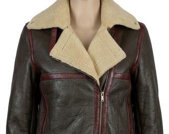 May include: A dark brown leather jacket featuring a cream-colored shearling collar and lapels. The jacket includes deep red leather trim accents along the seams, a silver-toned metal zipper closure, and a matching leather belt at the waist.