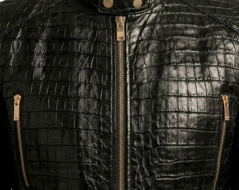 May include: A black jacket featuring a textured crocodile-skin pattern, a central gold-toned zipper, and two vertical gold-toned zippered pockets on the chest. The collar includes a snap-button closure with two gold-toned metal buttons.