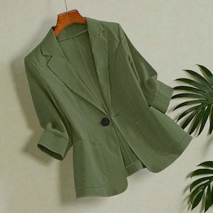 May include: A sage green blazer with three-quarter sleeves, a notched lapel, and a single black button closure. The blazer has two front pockets and is hanging on a wooden hanger.
