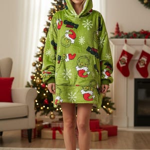 Puede incluir: Una manta con capucha verde con un dise&ntilde;o de Grinch y copos de nieve. La manta tiene un gran bolsillo delantero y una capucha forrada de blanco. La persona lleva zapatillas marrones. Un &aacute;rbol de Navidad y una chimenea est&aacute;n en el fondo.
