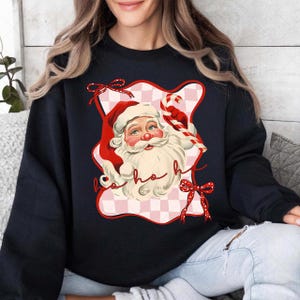 Könnte beinhalten: Schwarzes Sweatshirt mit einem Weihnachtsmann-Motiv. Das Design zeigt einen roten Hut, einen weißen Bart und eine Zuckerstange. Die Worte "ho ho ho" sind in einer verspielten Schriftart geschrieben. Das Weihnachtsmann-Motiv ist vor einem rosa-weiß karierten Hintergrund mit roten Schleifen platziert.