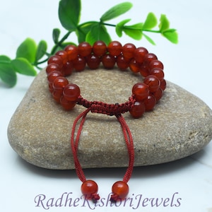 May include: A double-strand bracelet made of smooth, translucent, deep orange-red beads. The bracelet features a dark red adjustable cord closure with two dangling beads at the ends. The text RadheKishoriJewels is printed at the bottom.
