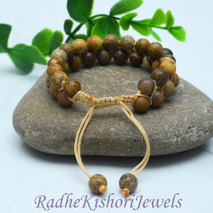May include: A double-strand bracelet featuring round, earth-toned jasper beads in shades of tan, brown, and beige. The bracelet includes a beige woven cord with an adjustable sliding knot closure and two dangling beads at the ends of the cords.