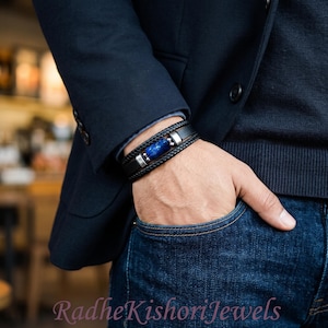 May include: A multi-strand black leather bracelet featuring a central cylindrical blue stone bead with metallic silver accents. The bracelet is worn on a wrist, layered with a dark navy blue blazer and a dark sweater, with the text RadheKishoriJewels.