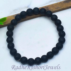 May include: A circular bracelet composed of numerous matte black lava stone beads with a porous, textured surface. The beads are strung together on an elastic cord. The text RadheKishoriJewels is printed in a dark font at the bottom of the image.