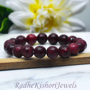 May include: A stretch bracelet composed of round, faceted beads in deep shades of burgundy and dark red with black mottled patterns. The bracelet is positioned on a wooden strip, and the text RadheKishoriJewels is printed at the bottom.