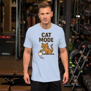 Könnte beinhalten: Hellblaues T-Shirt mit dem Schriftzug "CAT MODE" und einer Katzenillustration. Die Katze ist orange und braun und greift nach einer fallenden weißen Tasse. Das kurzärmelige T-Shirt wird von einer Person in einem Fitnessstudio getragen.