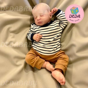 Op de afbeelding: Een realistische babypop met een zwart-wit gestreept shirt met lange mouwen en een bruine broek. De pop ligt op een beige ondergrond. De pop heeft gesloten ogen en een vredige uitdrukking. De afbeelding bevat een roze logo met "DEDA BABY SHOP".
