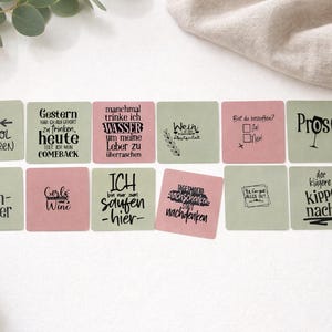 May include: A set of square coasters in pink and green, each with black text. The coasters display phrases such as "Girls Wine" and "Prost!" and are arranged on a white surface. Each coaster is approximately 10 cm x 10 cm.