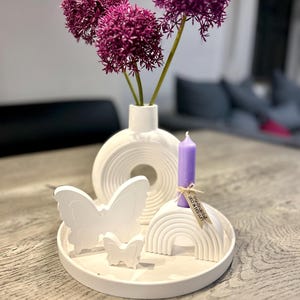 May include: A white ceramic vase holds three purple allium flowers. A white tray displays two butterfly figurines, a rainbow-shaped sculpture, and a purple candle. The arrangement is a decorative centerpiece.