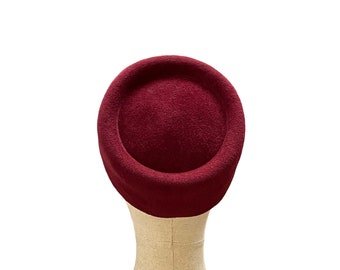 May include: A burgundy felt hat with a round crown and a wide brim. The hat is sitting on a fabric-covered mannequin head.