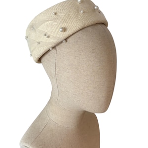 May include: A cream-coloured pillbox hat adorned with faux pearls. The hat, made from woven fabric, is displayed on a mannequin head. The design is simple, with a rounded crown and a slightly curved brim. Suitable for special occasions, it is a classic accessory.