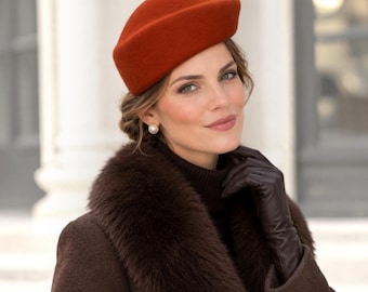 May include: A burnt orange felt pillbox hat with a structured, rounded crown. The hat is paired with a dark brown coat featuring a thick, plush faux fur collar, a dark brown turtleneck sweater, and dark brown leather gloves.