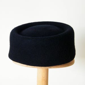 May include: A dark navy blue hat with a cylindrical crown and a flat top. The hat is made of a soft, textured material and sits on a wooden stand. The hat's design is simple and classic.