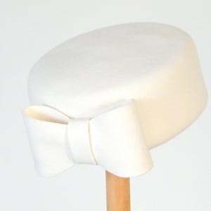 May include: A white felt hat with a large bow on the side. The hat is sitting on a wooden stand.