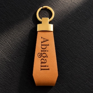 May include: A tan leather keychain featuring a gold-toned metal ring and a rectangular connector. The leather strap is embossed vertically with the name Abigail in a dark brown serif font, centered on the smooth surface of the material.