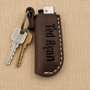 May include: A dark brown leather lighter case with visible light-colored stitching and the name Ted Ryan engraved vertically on the front. A metal key ring is attached to the top of the case, holding two metal keys, one silver and one brass.