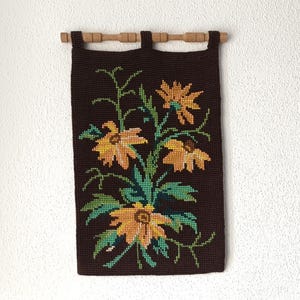 Peut inclure: Une tenture murale rectangulaire marron foncé avec un motif floral. Les fleurs sont jaunes et orange avec des feuilles et des tiges vertes. Elle est fixée à une tringle en bois.