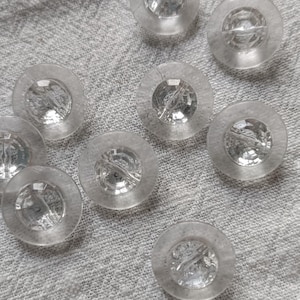 Op de afbeelding: Een verzameling van negen heldere, ronde knopen met een facetgeslepen, juweelachtig midden. De knopen zijn gerangschikt op een textiel, lichtgrijze stof. Elke knoop heeft een licht verhoogde rand en een klein gaatje in het midden.