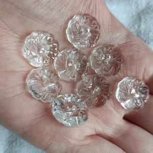 Puede incluir: Una colección de cuentas de vidrio transparentes en forma de flor. Cada cuenta tiene un diseño floral detallado y se sostiene en una mano. Las cuentas son transparentes y parecen ser de cristal o vidrio.