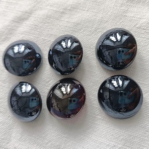 Puede incluir: Seis piedras esféricas pulidas, de color gris oscuro, con una superficie reflectante. Las piedras están dispuestas sobre una tela blanca texturizada. Las piedras tienen un aspecto liso y brillante.