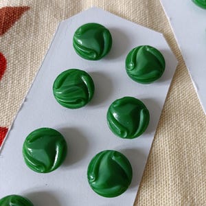 Puede incluir: Primer plano de varios botones redondos verdes con un diseño en espiral. Los botones están dispuestos sobre etiquetas de papel blanco, con una tela beige texturizada en el fondo. Los botones tienen un acabado brillante.