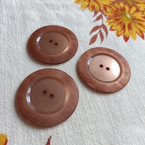 以下が含まれることがあります： それぞれ2つの穴がある、3つの大きくて丸い茶色のボタン。ボタンは木のような質感で、黄色、オレンジ、茶色の花柄の明るい色の生地に配置されています。
