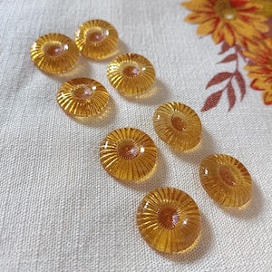 Peut inclure: Un ensemble de huit boutons en verre de couleur ambre. Chaque bouton présente une finition translucide avec un motif radial semblable à un rayon de soleil, composé de fines crêtes en relief partant du centre vers le bord extérieur.