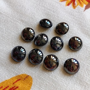 Puede incluir: Once piedras oscuras, brillantes y ovaladas dispuestas sobre una tela gris claro. Las piedras reflejan la luz, creando reflejos y sombras. Un estampado floral naranja y amarillo es visible en la esquina superior derecha e inferior izquierda.