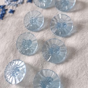 May include: A collection of eight light blue, round buttons. Each button has a flower design in the center and a ridged, sunburst pattern around the edge. The buttons are on a white, textured fabric.