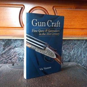 Könnte beinhalten: Ein Buch mit dem Titel "Gun Craft" mit blauem Einband. Das Cover zeigt ein detailliertes Bild einer Schusswaffe. Der Untertitel lautet "Fine Guns & Gunmakers in the 21st Century" von Vic Venters.
