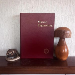 May include: A maroon book with the title "Marine Engineering" in gold lettering. A wooden mushroom ornament and a small animal figurine sit on a wooden shelf.